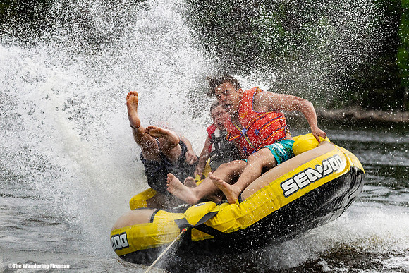 Three boys on a tube behind a boat in a huge water spray